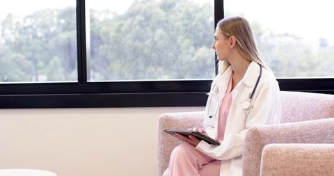 Female clinician in pink scrubs and white coat holding tablet gazing out clinic window