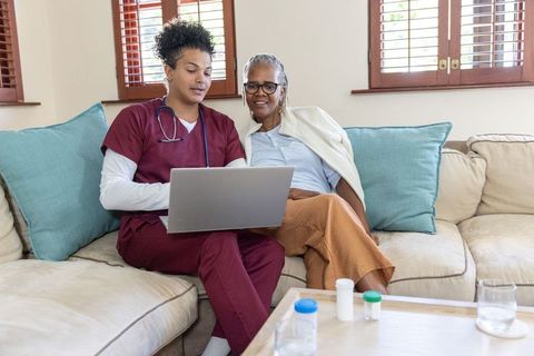 Home care nurse using laptop with senior woman on sofa reviewing medication together at home