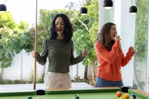 Female Friends Singing and Playing Pool in Sunlit Home Game Room with Glass Wall