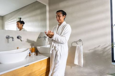 Asian woman applying moisturizer in white robe at modern minimalist bathroom vanity