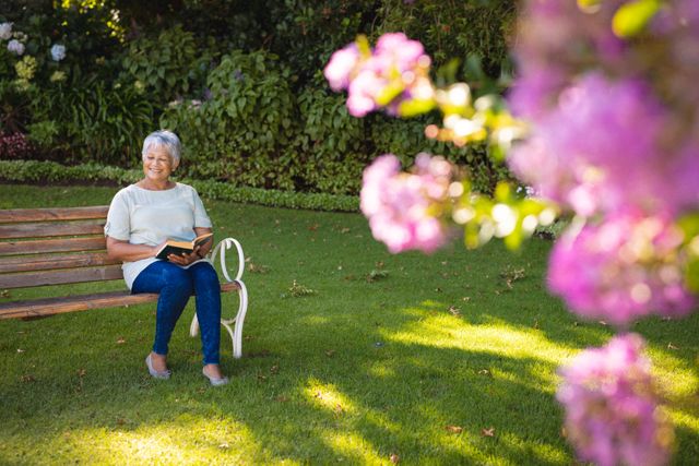 Senior woman enjoying a peaceful moment reading a book on a park bench surrounded by greenery and flowers. Ideal for use in articles or advertisements related to retirement, leisure activities, healthy lifestyle, and outdoor hobbies.