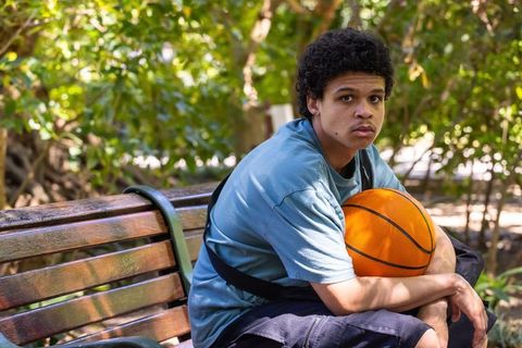 African american teen sitting on bench holding basketball, pensive portrait in park