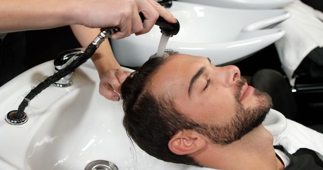 A young Caucasian man is getting his hair washed at a salon by a professional, with copy space. Capturing a moment of relaxation, the image reflects a common service provided in hair care establishments.