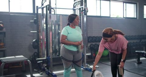 Diverse women chatting and hydrating after workout near squat rack and dumbbells