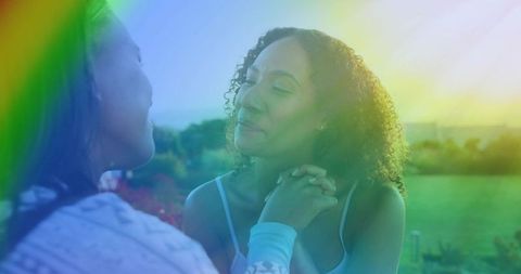 Two women holding hands in sunlit garden with rainbow lens flare, intimate warmth