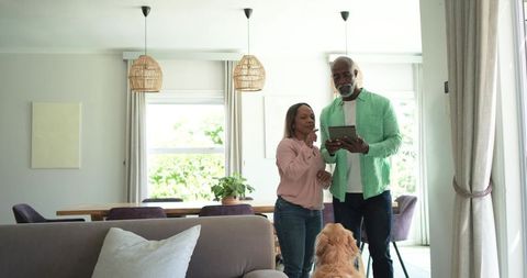 Senior couple standing using tablet with golden retriever in bright modern living room