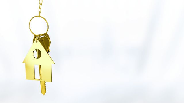 The golden house-shaped key dangles elegantly from a thin chain against a neutral white background, symbolizing concepts like home ownership, real estate investment, or security in property. This image can be used in real estate advertisements, promotional materials for housing developments, or articles related to home purchasing decisions. Its minimal design allows easy overlay of text in marketing applications.