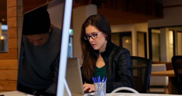 Young woman wearing glasses concentrating on work on her laptop while a male colleague stands next to her looking at the same device. Modern office setting with contemporary design. Ideal for illustrating concepts of teamwork, startups, professional work environments, and collaborative efforts.