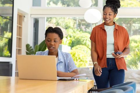 African american teen doing homework on laptop while mother observing with phone, workspace