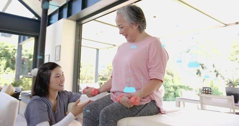 Senior woman lifting pink dumbbells on bench while caregiver guiding home exercise routine
