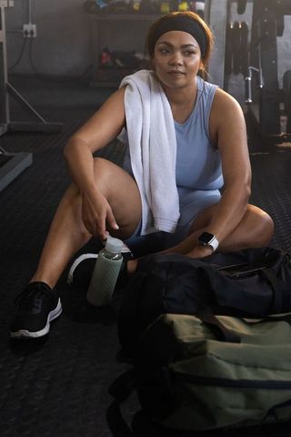 African american woman resting after workout holding water bottle and towel in gym with smartwatch