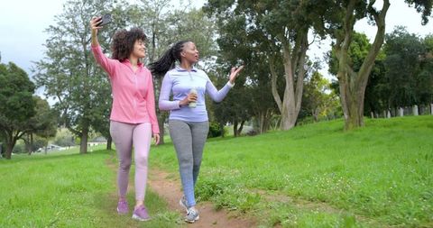 Women walking in park holding smartphone and water bottle during outdoor fitness walk