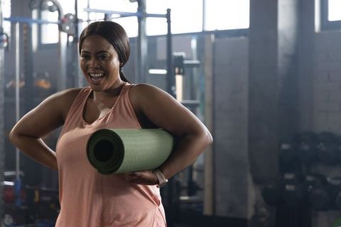 African american woman smiling and holding yoga mat in industrial gym wearing pink tank