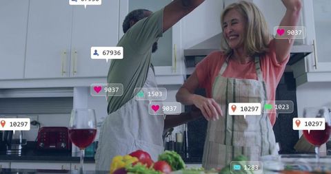 Mature couple dancing in kitchen while preparing vegetables with wine and social media icons