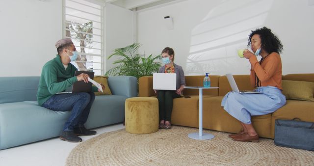 Biracial female and male colleagues in face masks sitting on sofas with computers and talking - Download Free Stock Photos Pikwizard.com