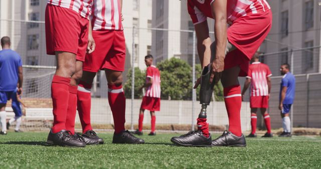 Soccer players in red uniforms preparing for match. Feature player adjusting prosthetic leg while interacting with teammates. Others in background wearing blue uniforms. Ideal for use in sports competition, disability awareness, team motivation, and athletic inspiration contexts.