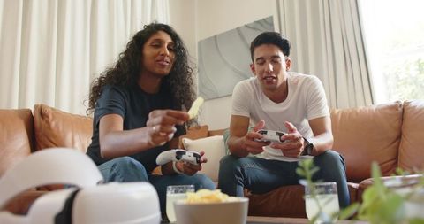 Young adults playing video games on couch with controllers and snacks casual living room