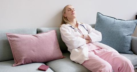 Female doctor resting on couch in staff lounge wearing white coat and pink scrubs