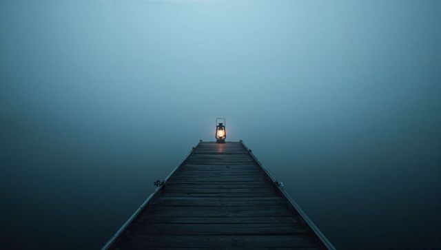 Wooden pier leading to lit lantern in dense fog, minimalist serenity, isolation and symmetry