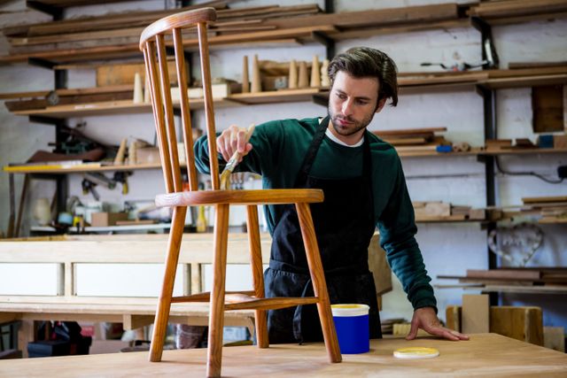 Carpenter varnishing a wooden chair in a workshop, showcasing craftsmanship and attention to detail. Ideal for use in articles about woodworking, DIY projects, small businesses, and artisan crafts. Can also be used in promotional materials for furniture makers and woodworking classes.