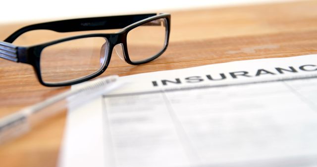 Glasses placed on a wooden desk beside a pen and an insurance document. This image can be used for illustrations related to business, financial services, insurance policies, paperwork, contractual agreements, and office environments.