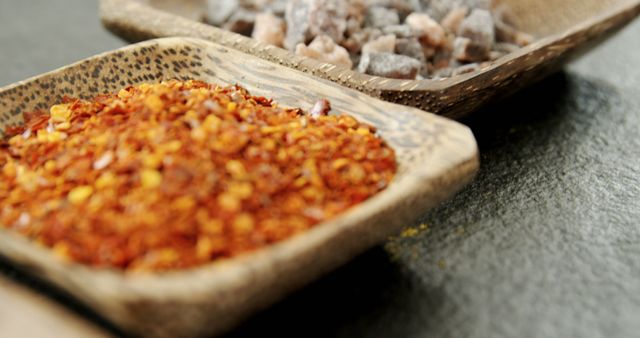 Two types of spices are presented in rustic bowls on a dark surface, with copy space. One bowl contains crushed red pepper flakes, while the other holds rock salt, suggesting a setting for cooking or seasoning food.