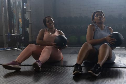 Two african american women performing core rotations holding medicine balls in gym