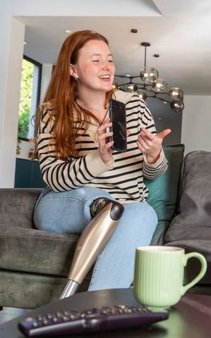 Red-haired woman smiling and showing prosthetic leg while using smartphone in modern living room