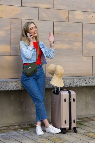 Young woman talking on phone waving beside suitcase in sunlit urban plaza casual denim travel style