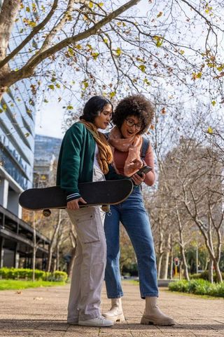 Diverse friends checking smartphone while holding skateboard on urban tree-lined path