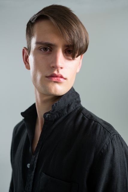 Man with an androgynous appearance and styled hair gazing at the camera. He is wearing a black shirt and standing against a grey background, creating a minimalistic and modern look. This can be used in fashion magazines, lifestyle blogs, or advertisements focusing on unique and modern styles.