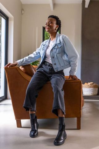 Joyful african american woman sitting on cognac armchair in modern sunlit living room