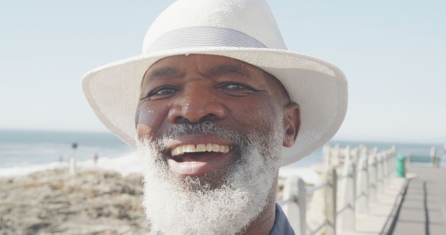 Elderly man smiling and enjoying a sunny day at the beachside, with a clear blue sky in the background and ocean waves in the distance. The man is wearing a white hat and appears joyful. This image can be used for promoting senior wellness retreats, happy lifestyle content for elderly individuals, or travel and vacation advertisements.