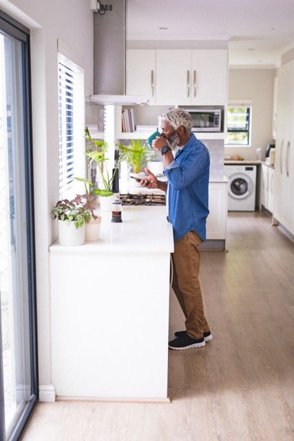Senior african american man drinking coffee while using smartphone in kitchen at home. unaltered, people, drink, technology and lifestyle concept.