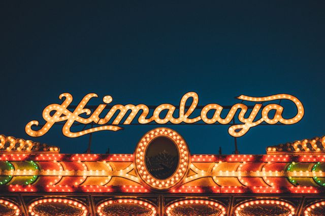 Bright, illuminated Himalaya ride sign glows at night, bringing nostalgia and excitement. Perfect for illustrating themes related to amusement parks, carnivals, festivals, entertainment, funfair rides. Highlights aesthetics of nighttime events and neon lights.