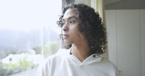 Non-binary person looking out window in white hoodie with curly hair, contemplative natural light po
