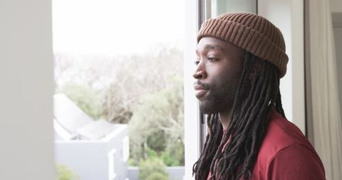 Young African American Man Gazing Out Apartment Window Wearing Beanie, Maroon Shirt, Contemplative