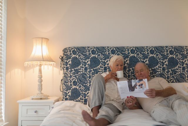Senior couple enjoying a peaceful morning in bed, reading the newspaper and drinking coffee. Ideal for topics on retirement, elderly lifestyle, morning routines, togetherness, and intimate moments at home. Perfect for advertisements, blogs, or articles focusing on senior living, home comfort, and family relationships.
