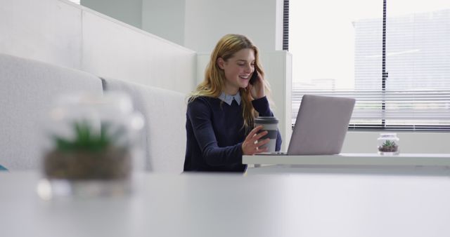 This image captures a young professional woman smiling while working on her laptop in a modern office environment. She holds a cup of coffee, suggesting a casual yet productive work atmosphere. Perfect for illustrating concepts related to business, career development, corporate culture, modern workplaces, remote work setups, and communication in professional settings.