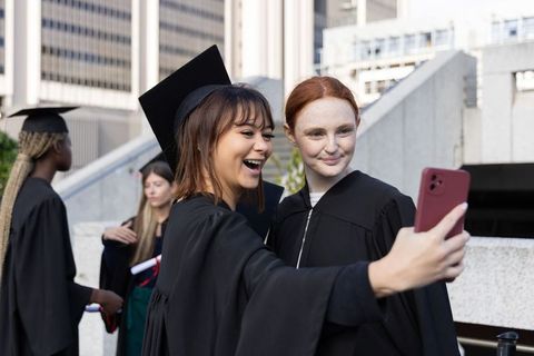 Graduating students taking selfie on urban campus courtyard wearing gowns