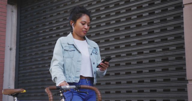 This image features a woman in an urban setting, walking with her bicycle while texting on her smartphone. She is casually dressed in a denim jacket and wearing earphones. This can be used to represent themes like urban lifestyle, connectivity, multitasking, and modern living.