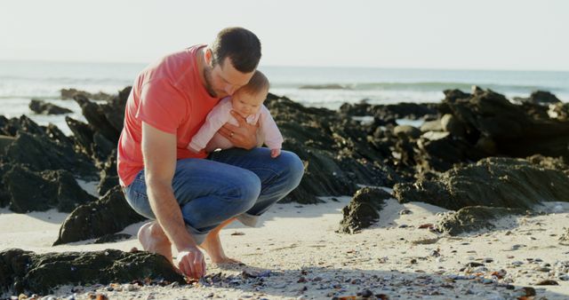 Caucasian father holding happy baby kneeling on sunny beach looking at shells, copy space. Fatherhood, care, wellbeing, vacations, nature and travel, unaltered.