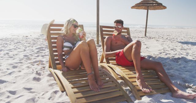 A couple is relaxing on wooden loungers under beach umbrellas on a sandy beach, enjoying refreshing drinks and the sunny weather. Perfect for illustrating travel brochures, tourism websites, summer vacation promotions, or lifestyle articles about relaxation and leisure activities.