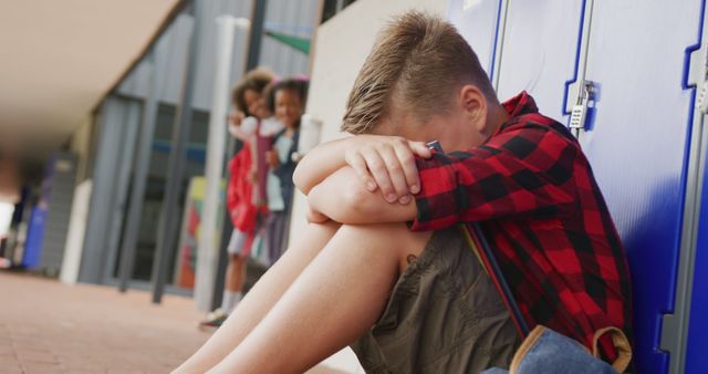Boy sitting alone by school lockers with his head down, while kids in the background mock him. This can be used for articles or presentations related to bullying, mental health awareness, educational materials on empathy and kindness, and social issues affecting children in schools.