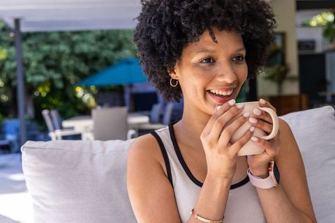 African american woman smiling while holding ceramic mug on shaded patio sofa