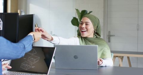 Smiling professional woman in green hijab exchanging usb drive with coworker at desk