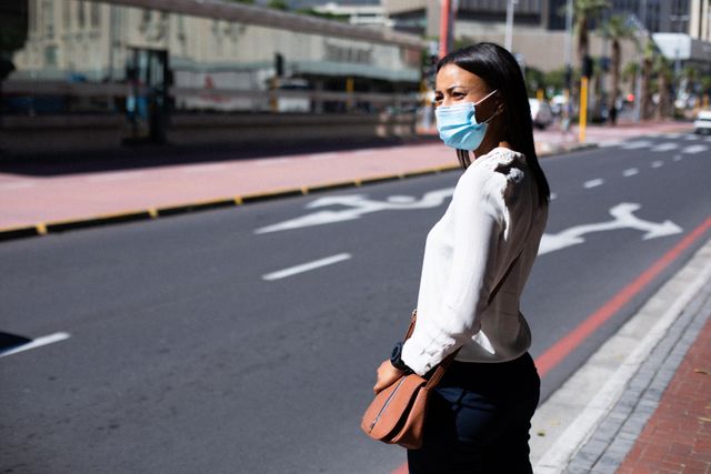 African American woman wearing a face mask standing on a city street. This image can be used for topics related to public health, safety measures during the coronavirus pandemic, urban lifestyle, and social distancing practices.