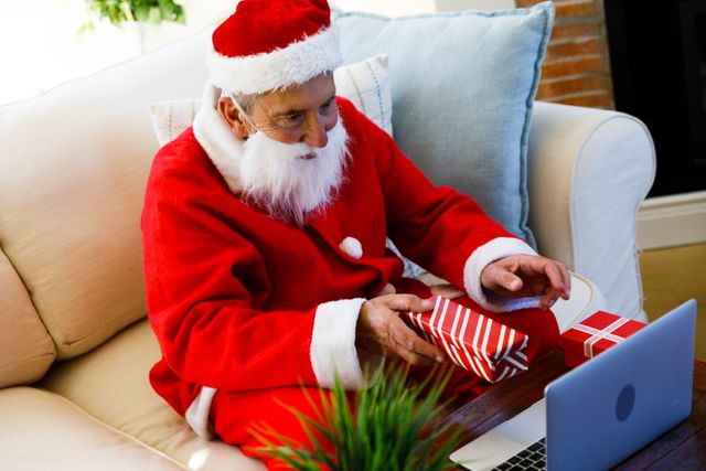 Elderly man dressed as Santa Claus holding a Christmas gift while on a video call using a laptop. Ideal for holiday-themed promotions, virtual celebration advertisements, and festive greeting cards. Highlights the blend of traditional holiday spirit with modern technology.