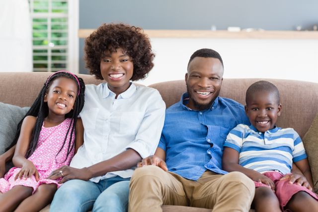 This image shows a happy African American family sitting together on a sofa in their living room. The parents and their two children are smiling, creating a warm and joyful atmosphere. This image can be used for family-oriented content, advertisements, parenting blogs, and articles about family life and togetherness.