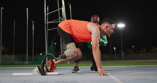 The image shows a male disabled athlete with a prosthetic leg preparing to sprint at night on a sports track, with a coach providing support. This image can be used for topics related to adaptive sports, motivation, coaching, and the strength of the human spirit.